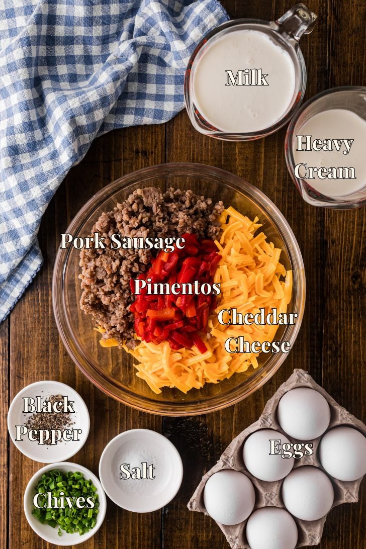 Fresh ingredients for pimento cheese sausage quiche including grated sharp cheddar, crumbled breakfast sausage, diced pimentos, eggs, whole milk, heavy cream, and a deep dish pie crust arranged on a countertop.