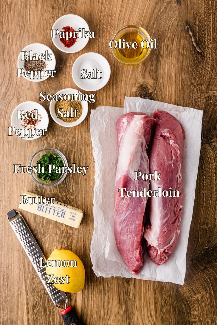 Ingredients for cast iron roasted pork tenderloin including pork, Dan-O's seasoning, paprika, butter, lemon, and fresh parsley on a wooden board.