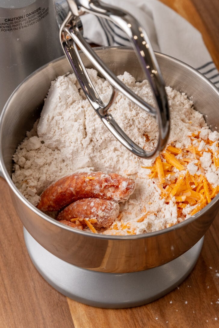 Overhead view of cheesy sausage balls 4 ingredients in metal mixing bowl: flour, shredded cheddar cheese, and raw sausage with electric mixe