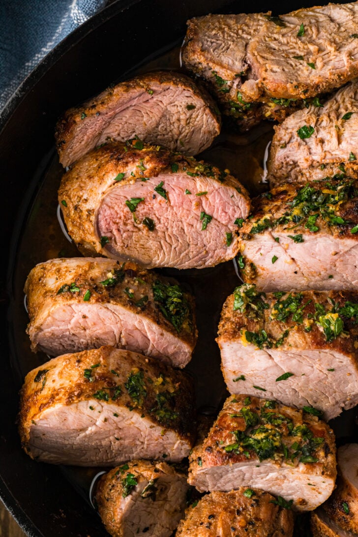 Sliced roasted pork tenderloin in a cast iron skillet with a golden herb butter crust and fresh parsley.