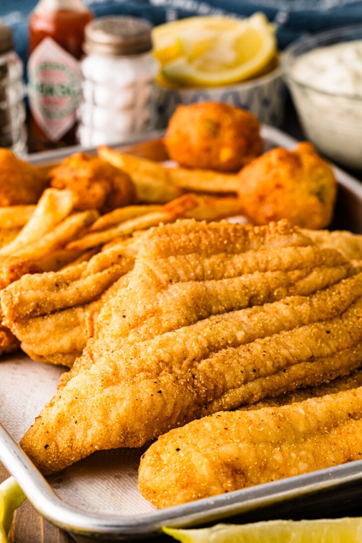 Southern fried catfish fillets on a tray with lemon wedges, hot sauce, tartar sauce, fries, and hush puppies.