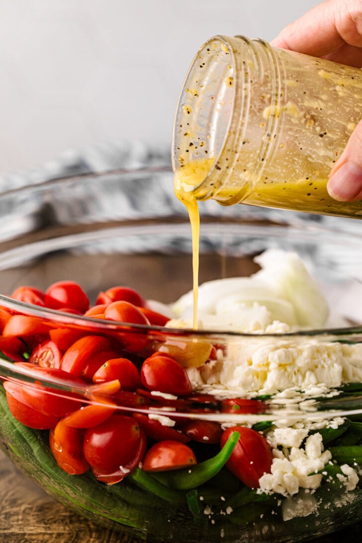 Lemon Dijon dressing being poured over green beans, grape tomatoes, onion, and feta for green bean salad.