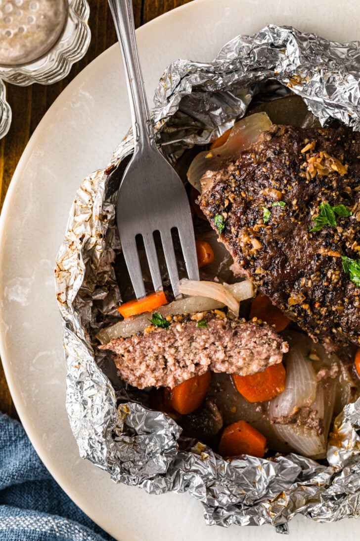 Fork cutting into a hamburger patty in a hobo dinner foil pack with potatoes, carrots, and onions.