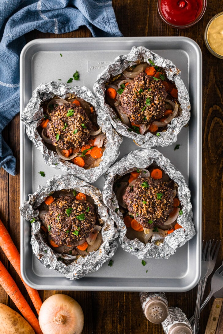 Four open hamburger hobo dinner foil packs on a baking sheet with beef patties and vegetables.