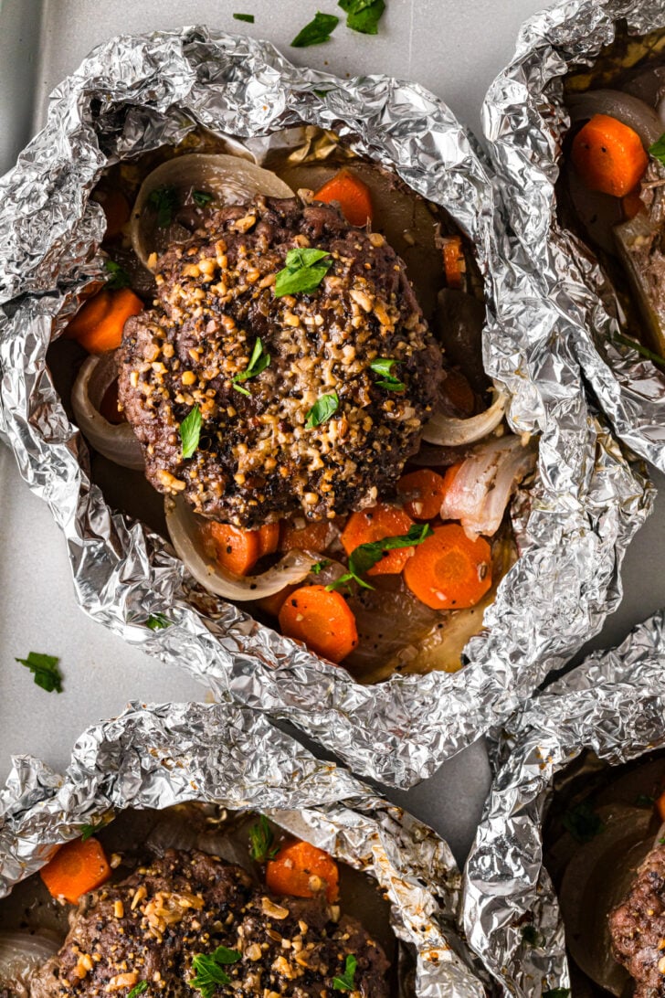 Hamburger hobo dinner foil packs with seasoned beef patties, potatoes, carrots, onions, and parsley.