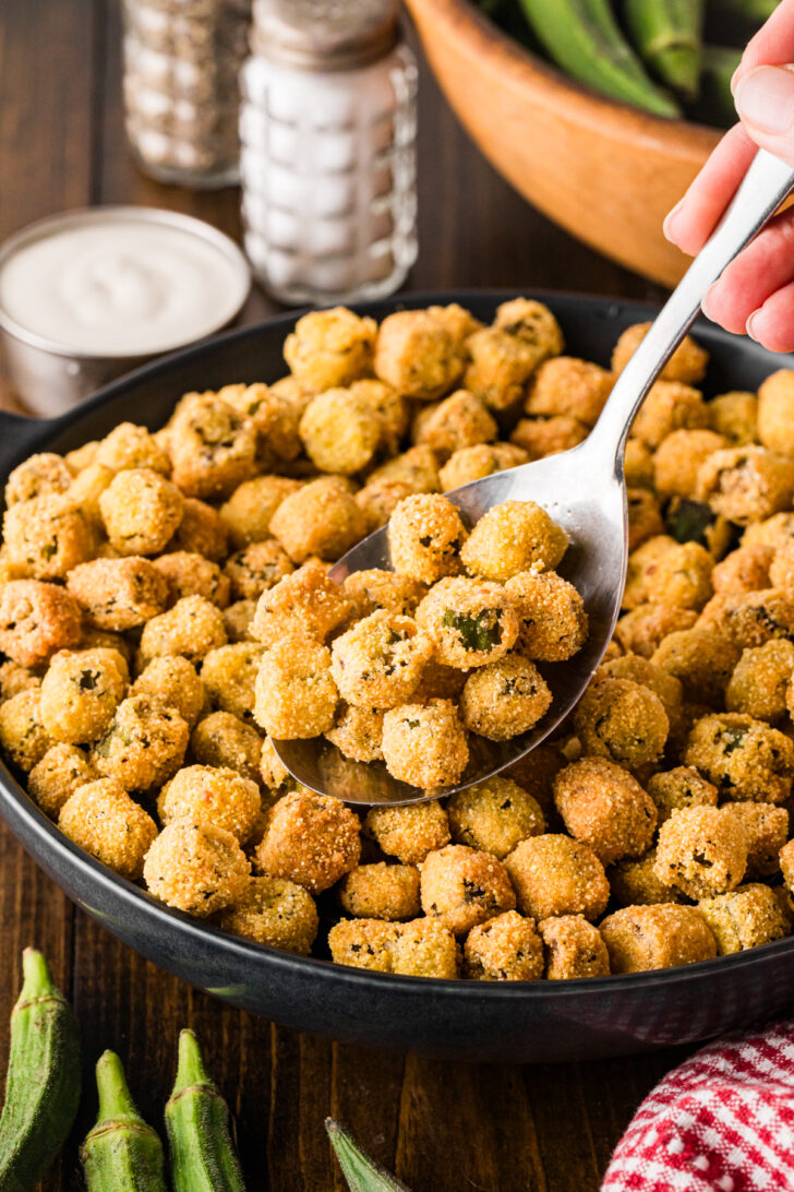 Hand scooping fried okra with a spoon from a black skillet.