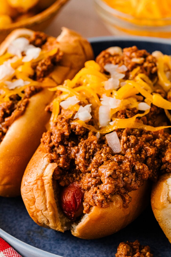 Chili cheese hot dogs topped with shredded cheddar cheese and diced onions on a blue plate.