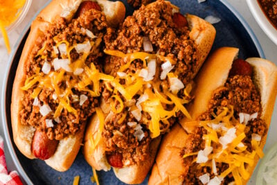Overhead view of three chili cheese dogs topped with cheddar cheese and diced onions on a blue plate.