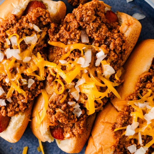 Overhead view of three chili cheese dogs topped with cheddar cheese and diced onions on a blue plate.
