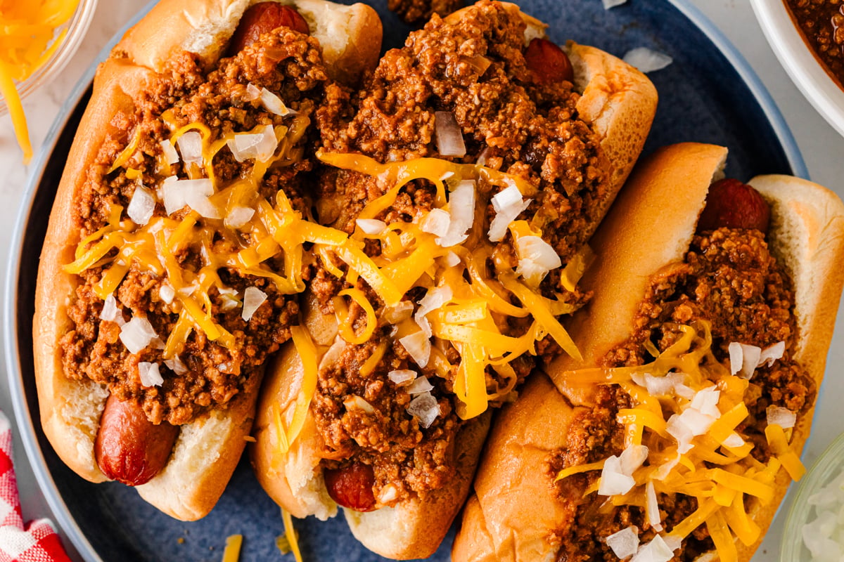 Overhead view of three chili cheese dogs topped with cheddar cheese and diced onions on a blue plate.