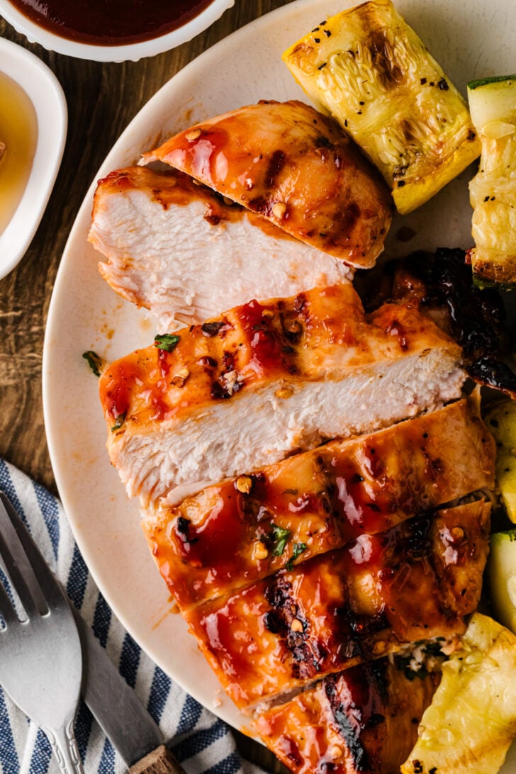 Sliced BBQ chicken breast on a plate with grilled squash and dipping sauce.
