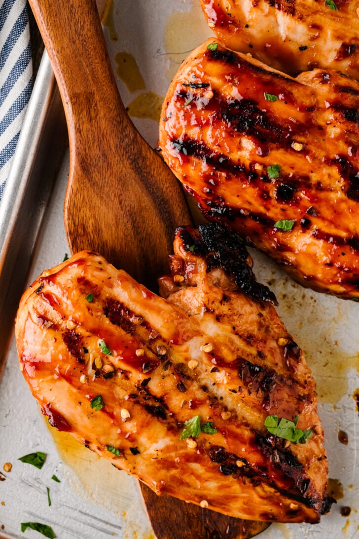 Grilled BBQ chicken breasts with a wooden spoon on a sheet pan.