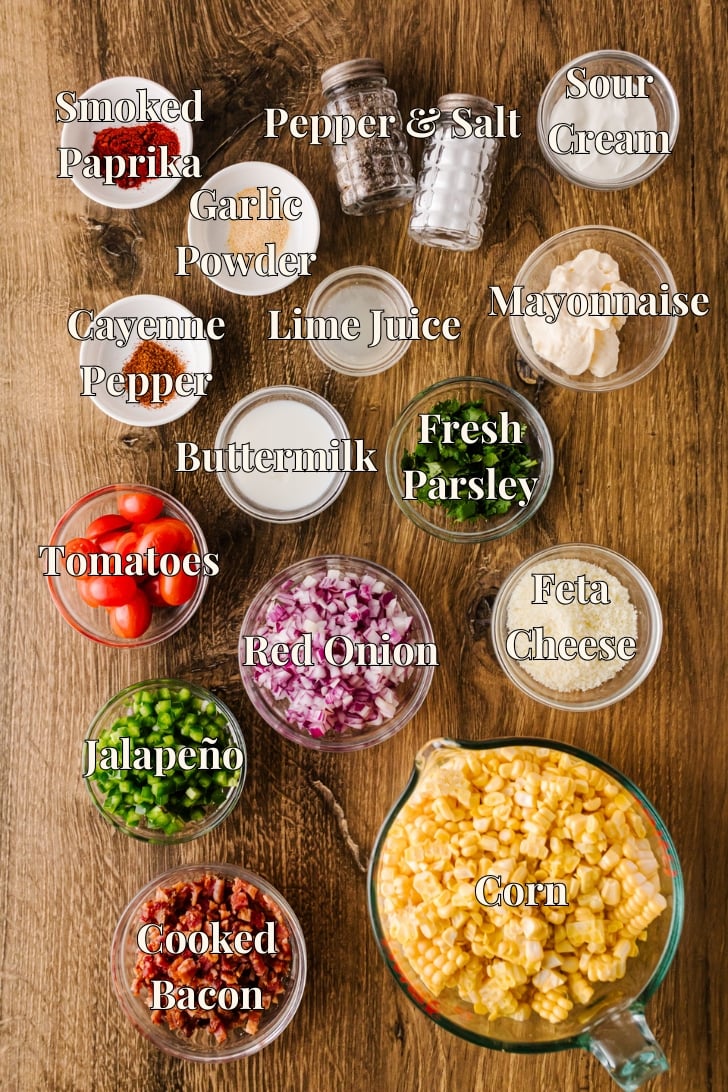 Ingredients for Southern corn salad arranged on a wooden table: corn, bacon, cherry tomatoes, red onion, jalapeño, feta, parsley, mayonnaise, sour cream, buttermilk, lime juice, smoked paprika, cayenne, garlic powder, salt, and pepper.