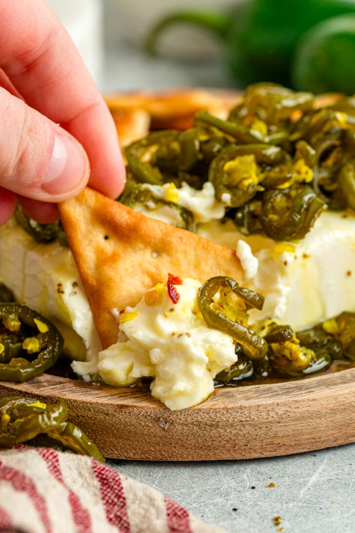 Hand holding a cracker topped with cream cheese and candied jalapeños, with fresh green jalapeños in the background.
