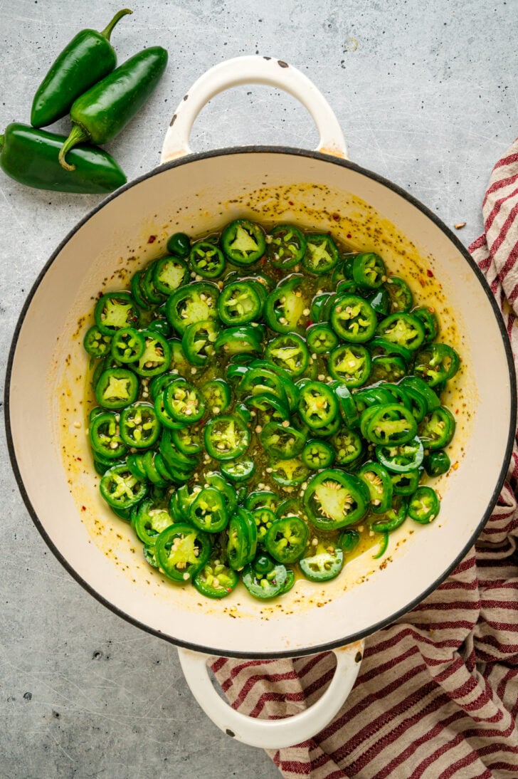 Open mason jar filled with candied jalapeño slices in sweet and spicy syrup beside a bowl of fresh green jalapeños.