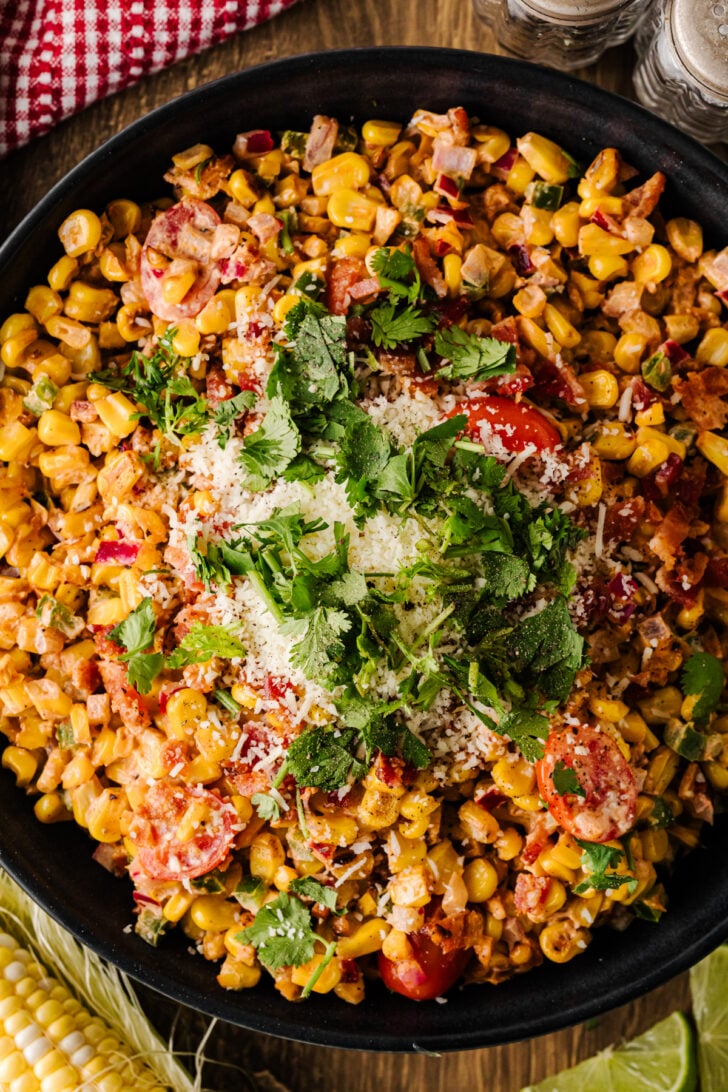 Southern corn salad served in a black oval bowl with sweet corn, bacon, cherry tomatoes, red onion, jalapeño, feta cheese, cilantro, lime wedges, and a serving spoon.
