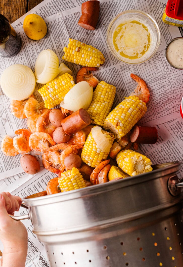Pouring shrimp, sausage, corn, and potatoes from a strainer pot onto a low country boil spread.