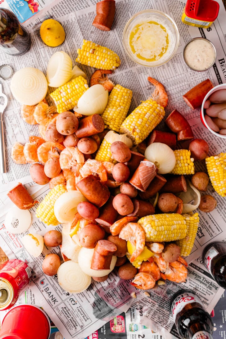 Overhead low country boil spread on newspaper with dipping sauces, drinks, and Old Bay seasoning.