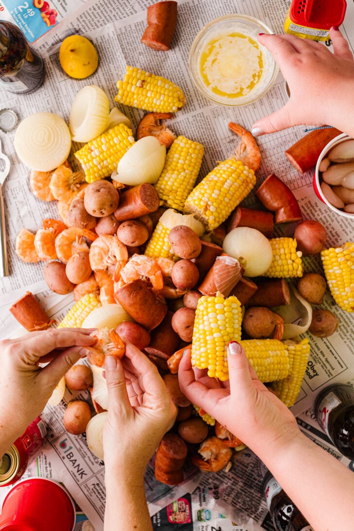 Hands digging into a low country boil spread on a newspaper-covered table with corn, shrimp, potatoes, and sausage.