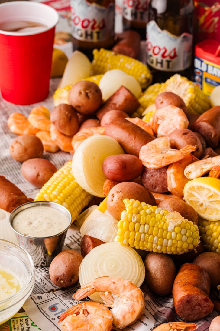 Low country boil spread on newspaper with shrimp, sausage, corn, potatoes, dipping sauces, and beer.