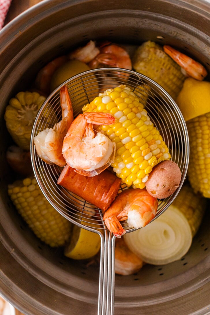 Slotted spoon scooping shrimp, corn, and sausage from a low country boil pot.