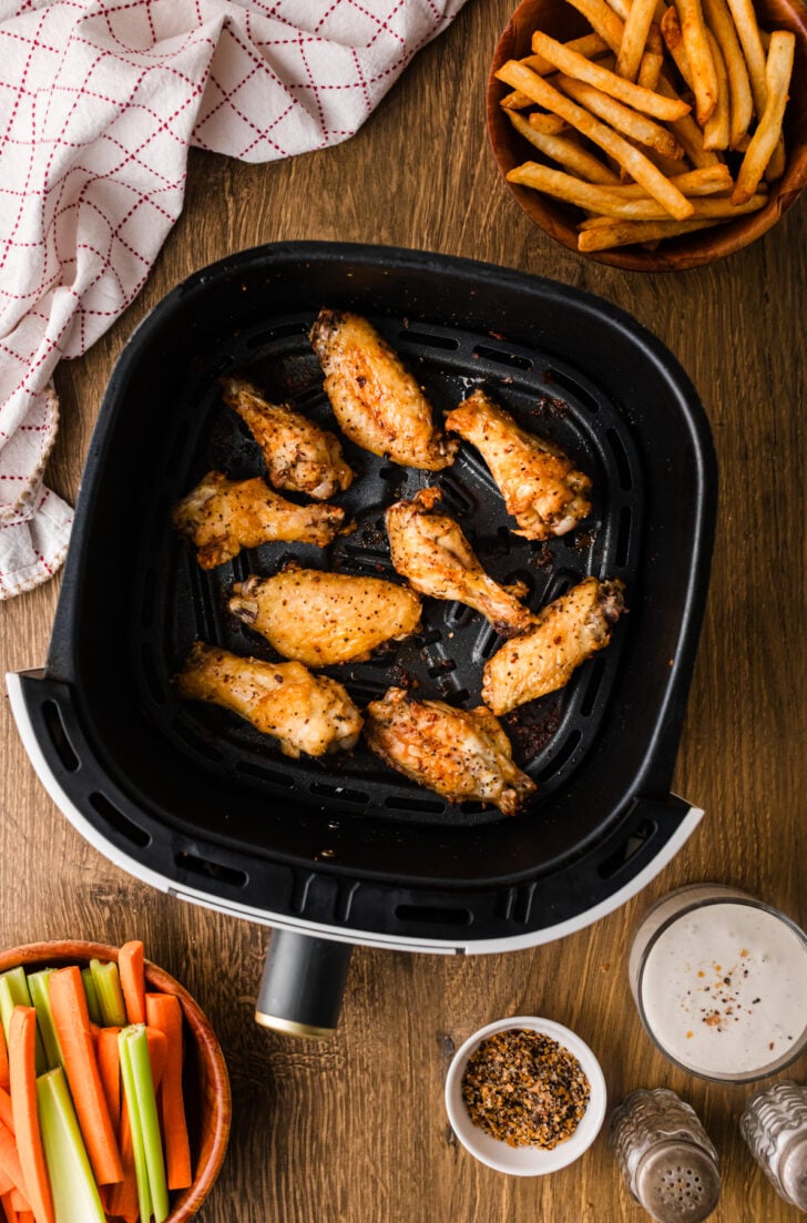 Seasoned chicken wings in a single layer in the air fryer basket, surrounded by carrot and celery sticks, fries, and dipping sauce.