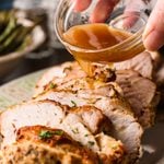 Hand pouring gravy from a glass bowl over sliced air fryer turkey breast on a serving platter.