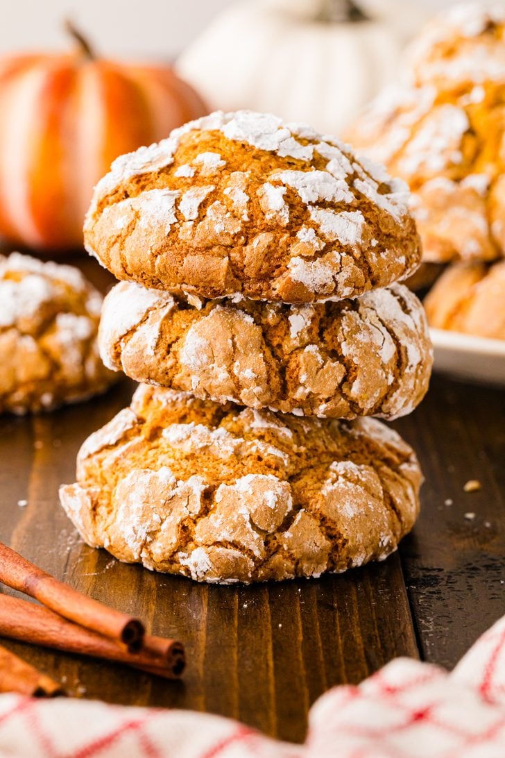 Stack of pumpkin crinkle cookies dusted with powdered sugar on a wooden table with pumpkins and cinnamon sticks.